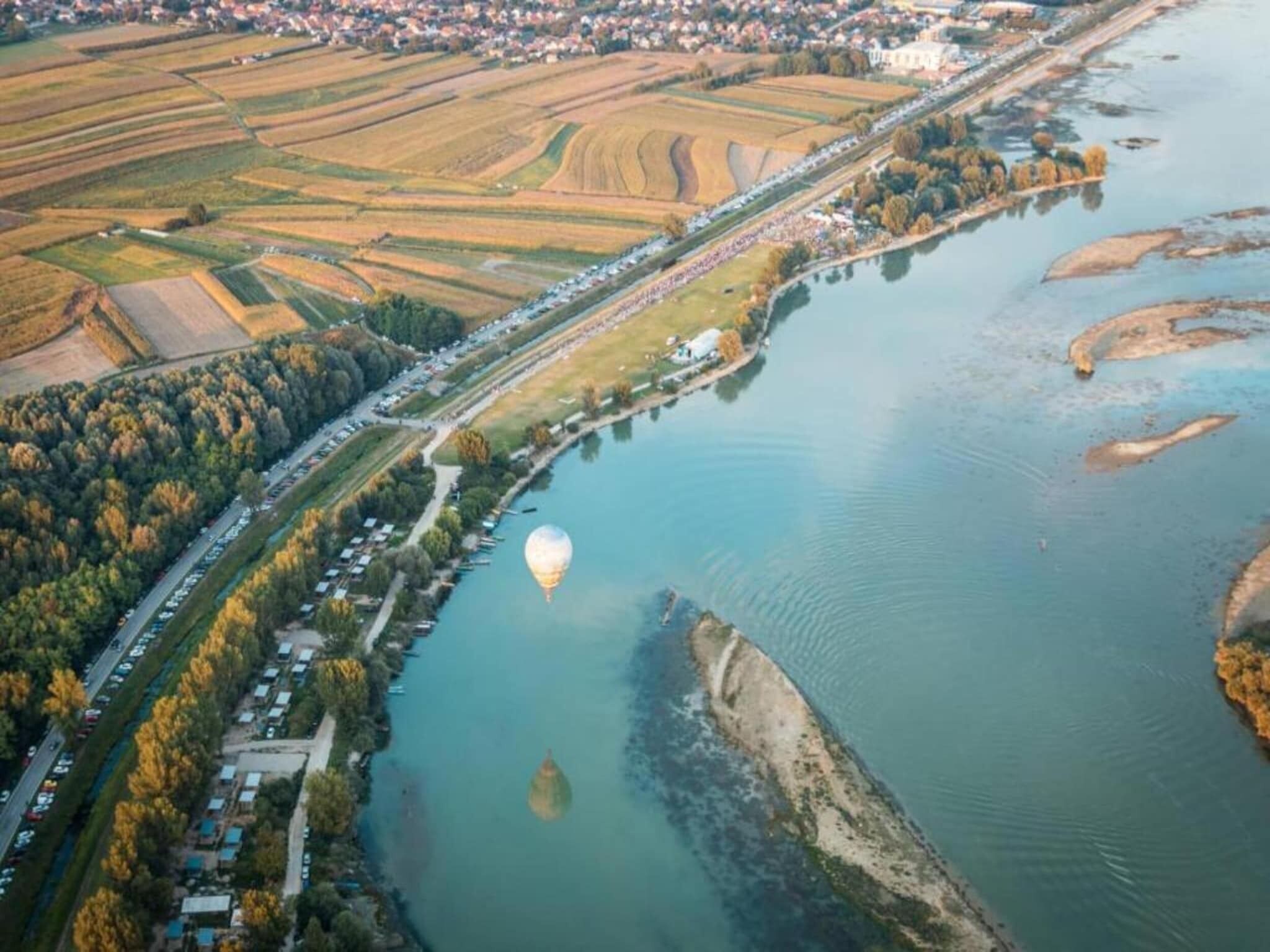 Une vue aérienne d’une rivière, avec une montgolfière au-dessus, entourée de champs et d’une petite ville.