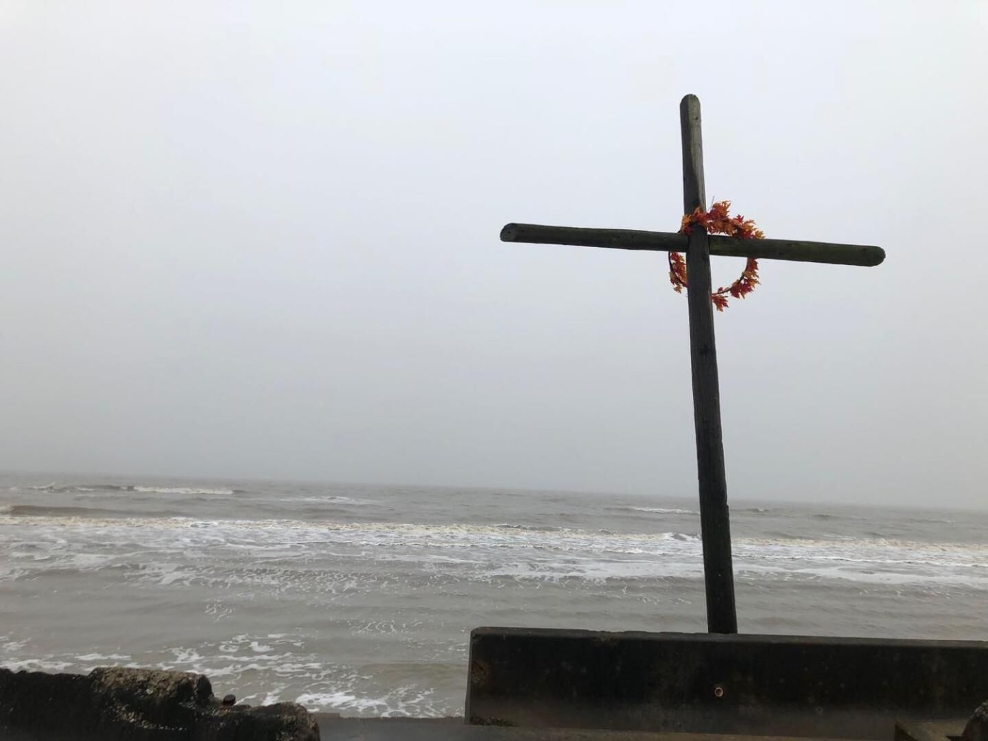 Une croix ornée d’une guirlande, placée sur un poteau au bord de la mer.