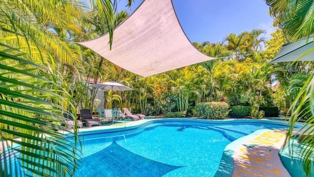 A swimming pool with a clear blue water, surrounded by palm trees and a shaded area. - Villas Verdes (Sámara)
