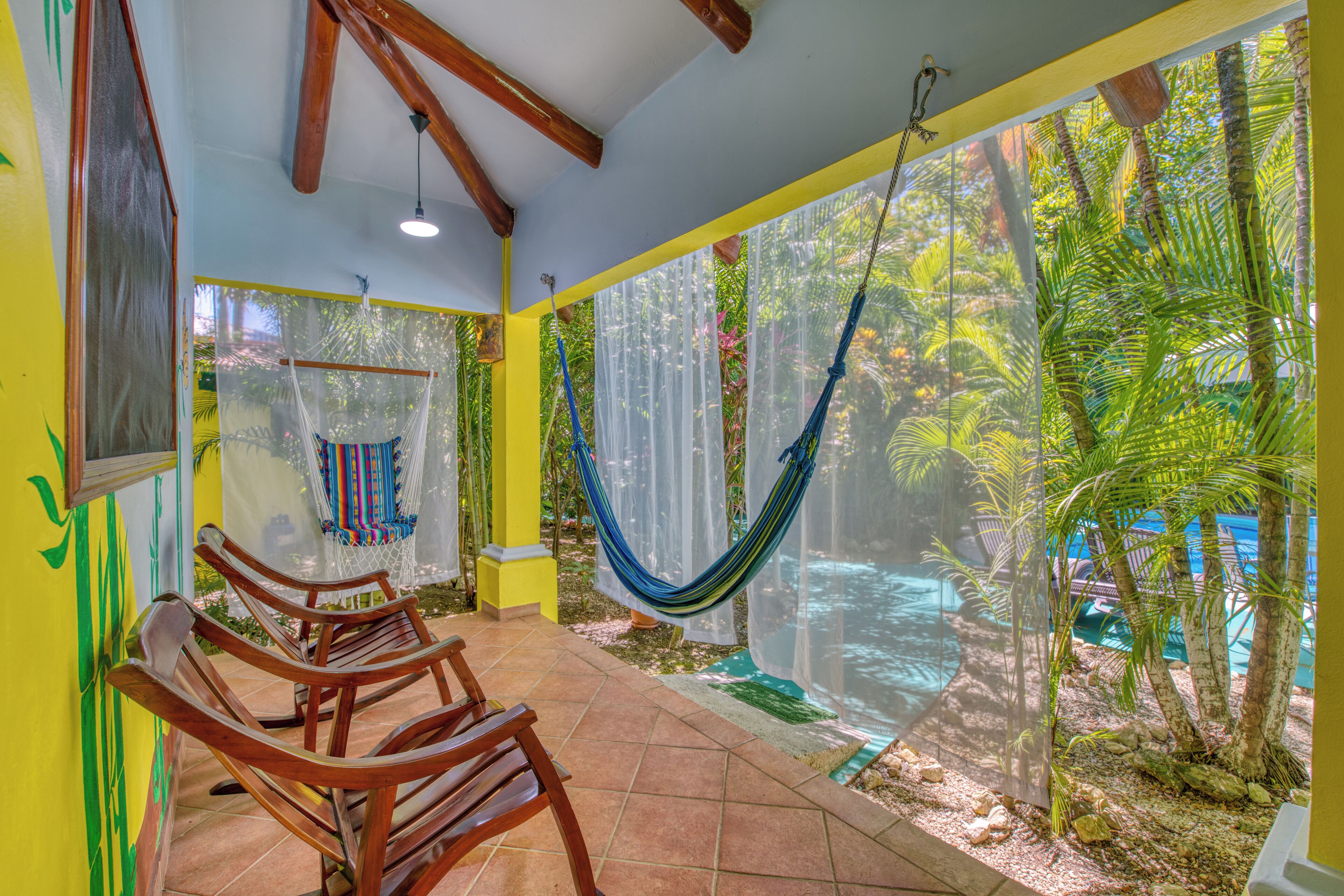 A room with wooden chairs, a hammock, and a view of a pool and palm trees.