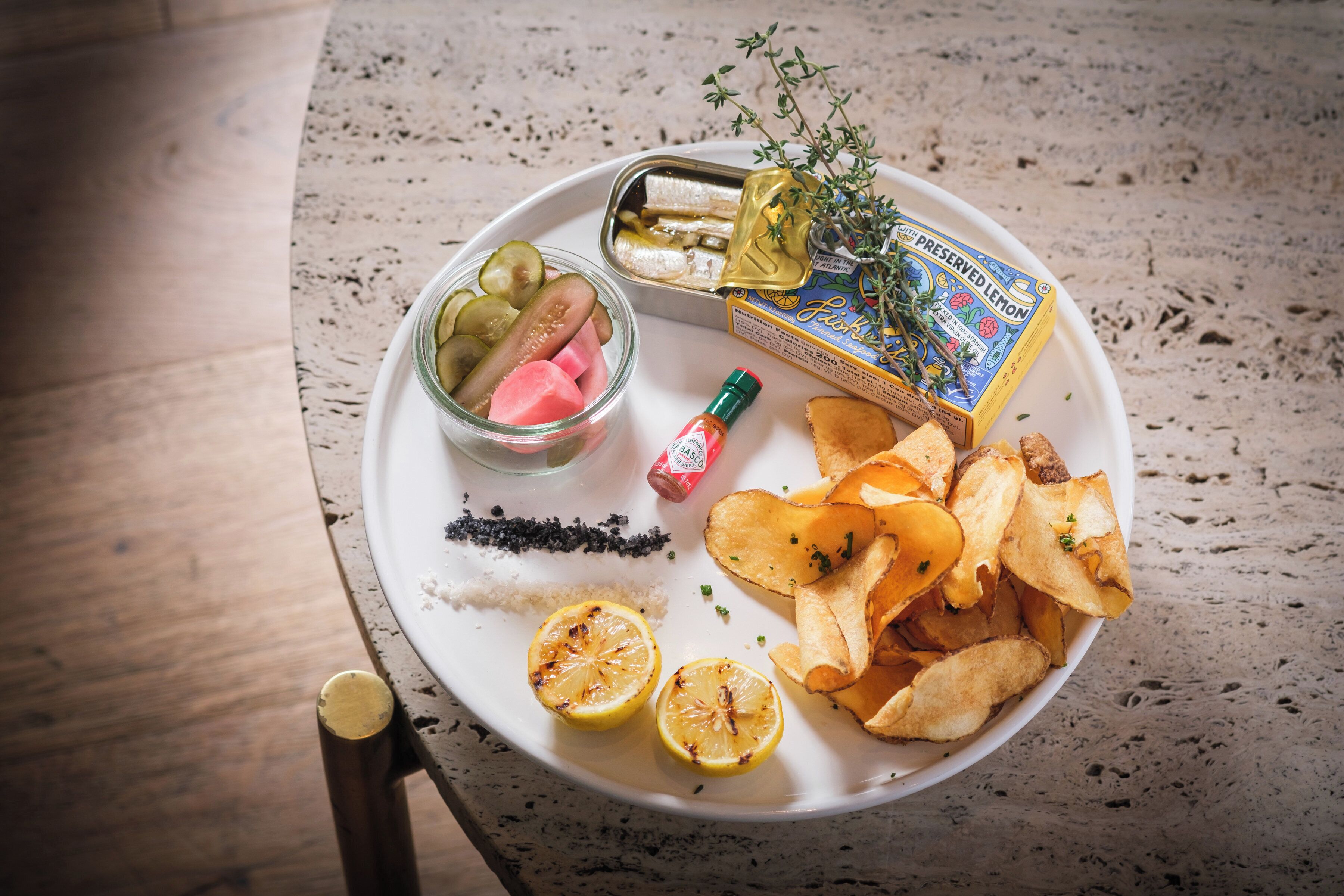 A plate with pickles, lemon slices, chips, and a bottle of sauce on a table.
