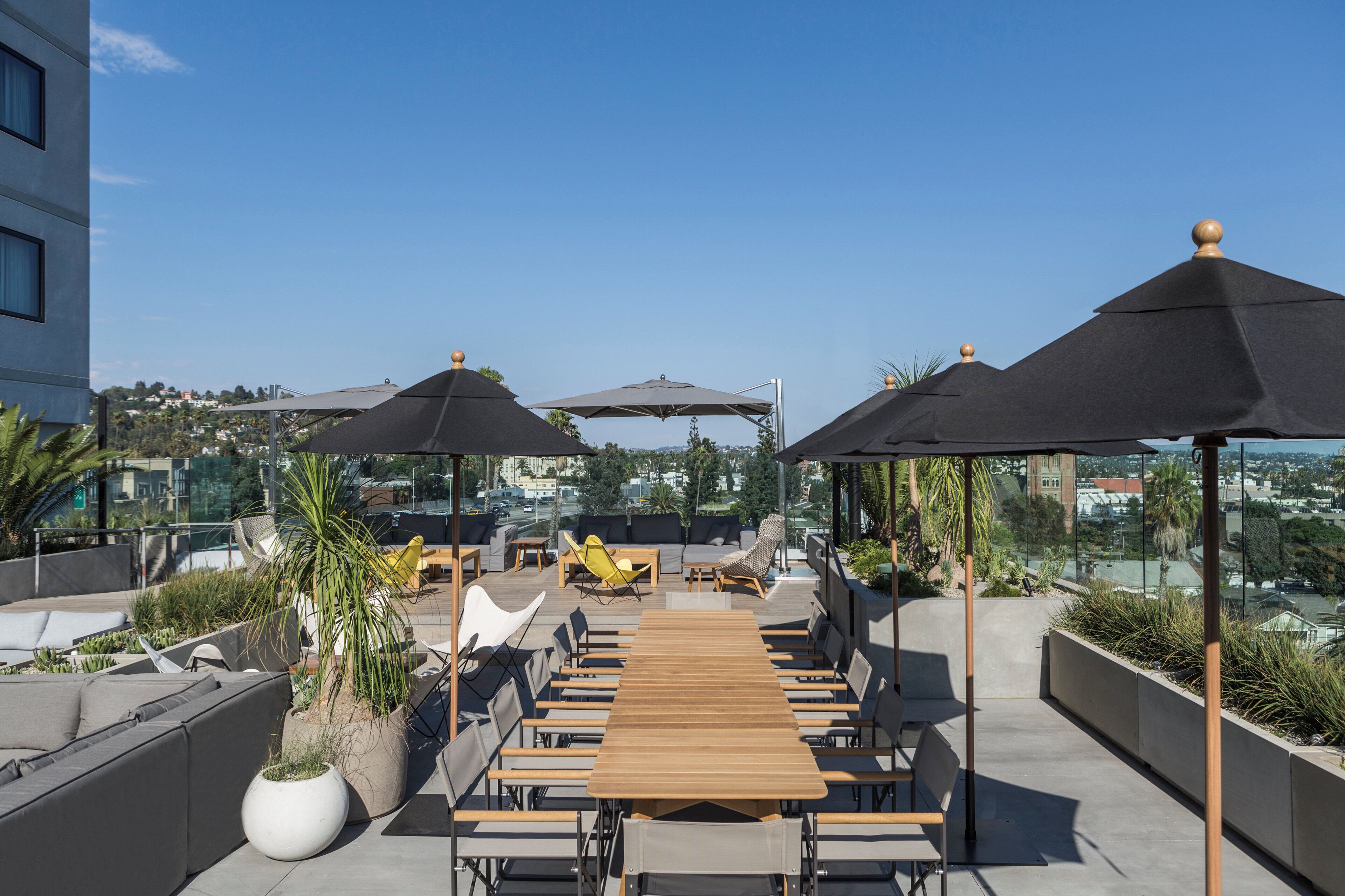 A rooftop terrace with a long wooden table, multiple chairs, and umbrellas providing shade.