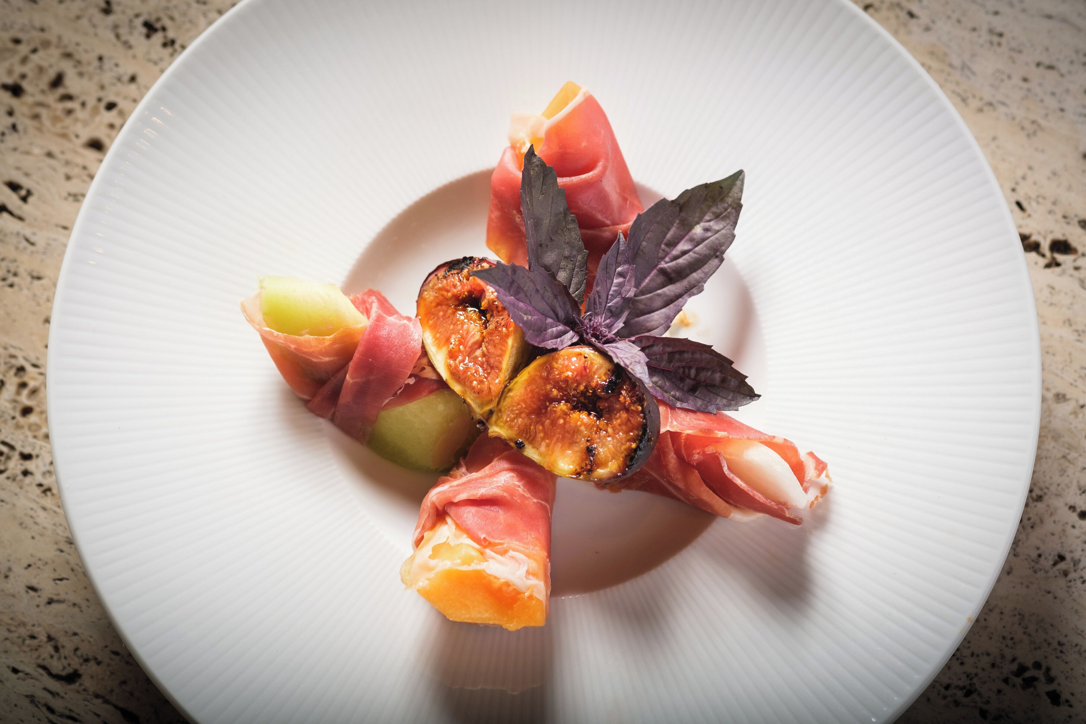 A white plate with a variety of food items including slices of cured meat, a piece of grilled fruit, and a decorative leaf.