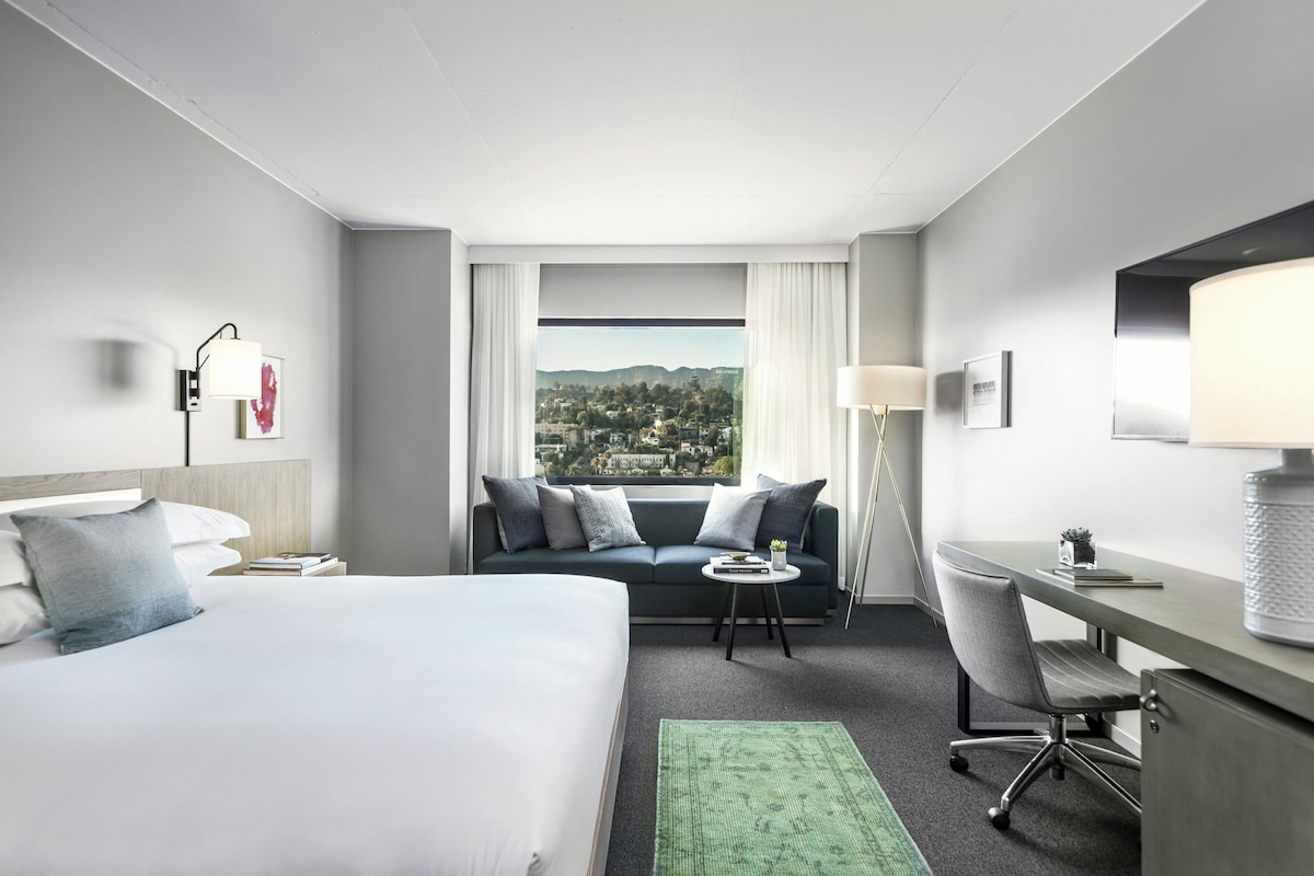 A hotel room with a bed, a sofa, a desk, a chair, a lamp, and a view of a cityscape.
