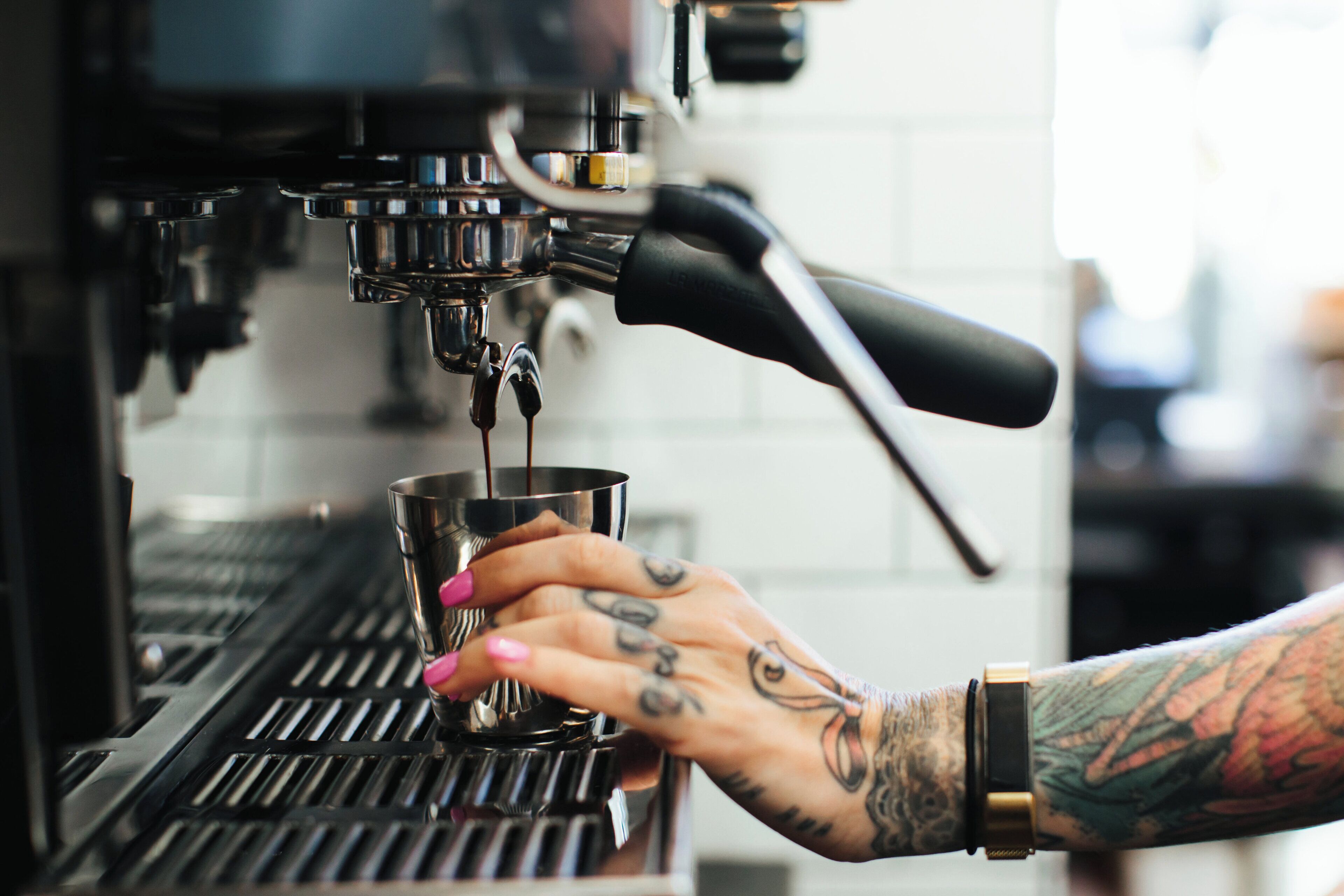 Une main tenant une tasse en métal sous une machine à café, et on peut voir le bras tatoué d’une personne.