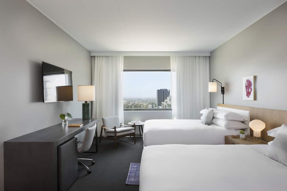 A hotel room with two beds, a desk, a chair, a lamp, and a view of the city.