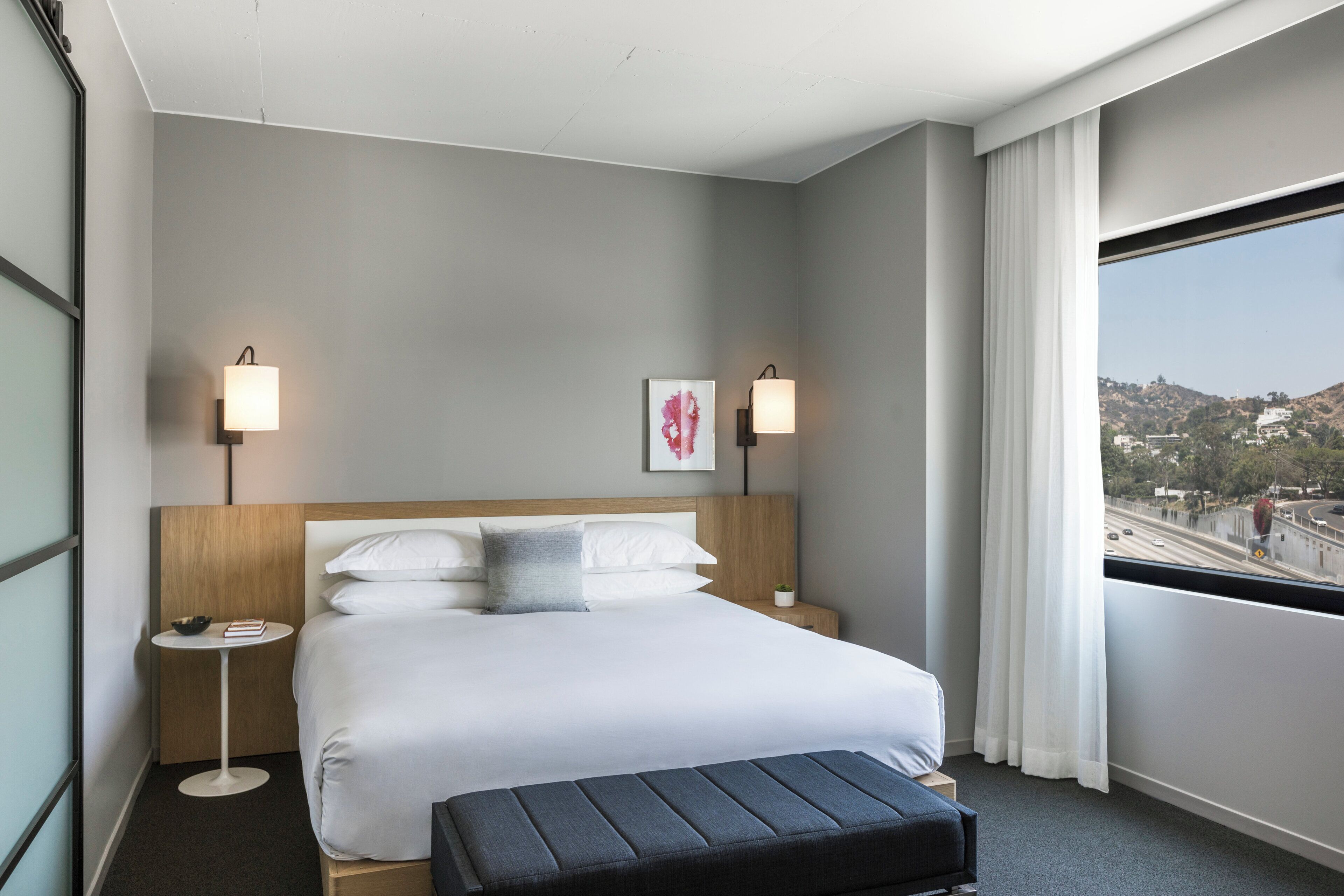 A hotel room with a bed, bedside tables, a small table, a bench, a window with a view, and wall-mounted lamps.