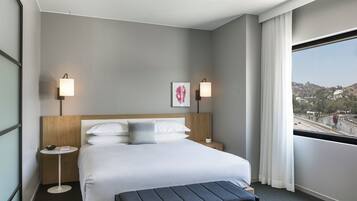 A hotel room with a bed, bedside tables, a small table, a bench, a window with a view, and wall-mounted lamps.
