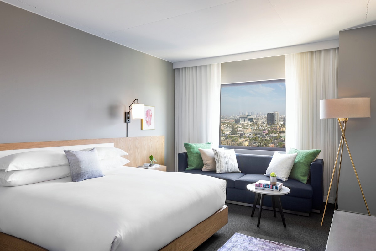 A hotel room with a bed, a sofa, a small table, a lamp, and a view of a cityscape.