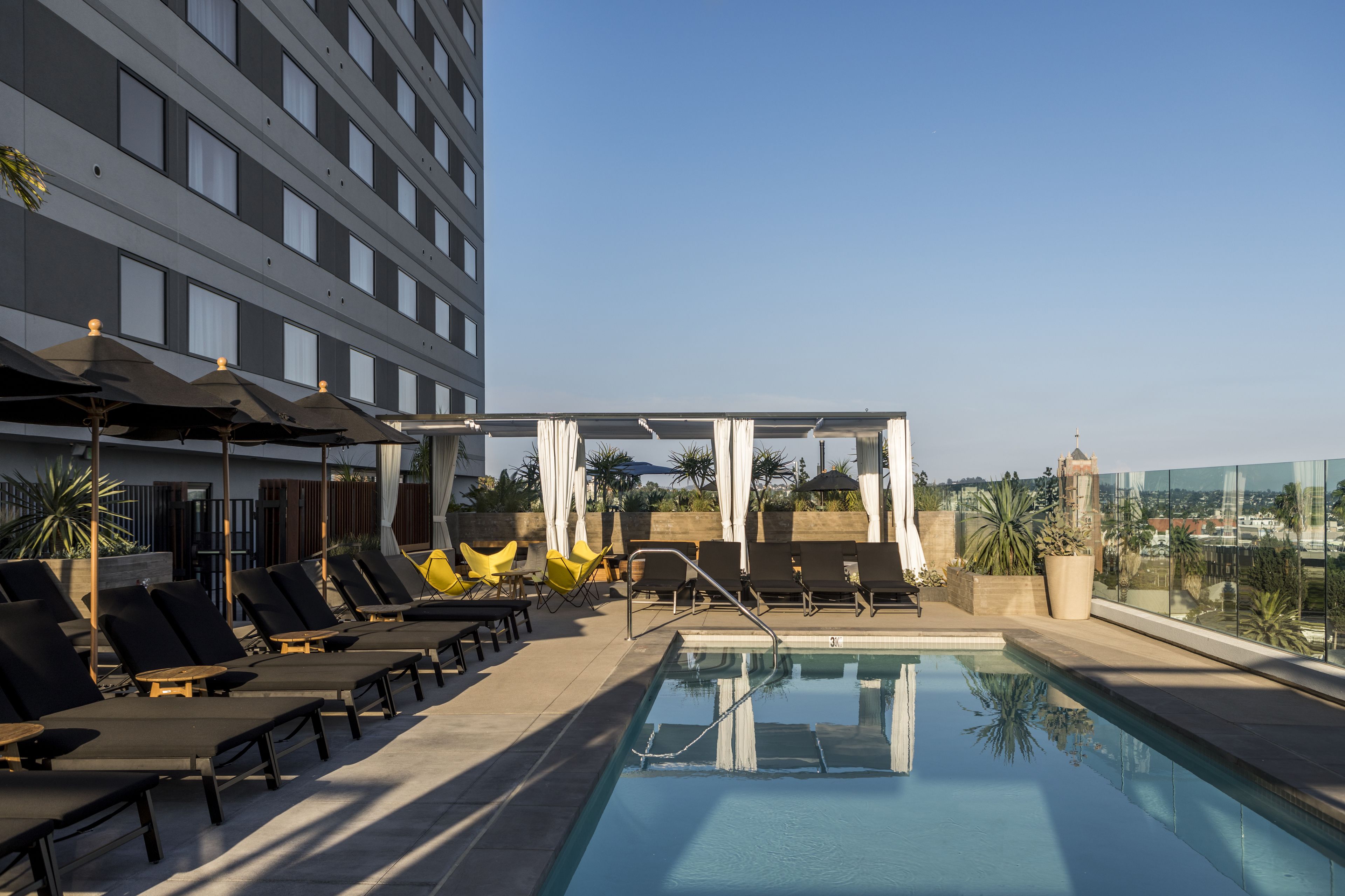 A rooftop pool with sun loungers, a glass balustrade, and a view of the cityscape.