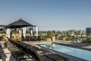 A rooftop pool area with lounge chairs and a gazebo, overlooking a cityscape.
