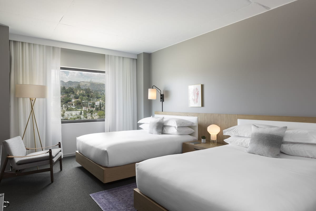 A hotel room with two beds, a chair, a bedside table with a lamp, and a view of a cityscape through large windows.