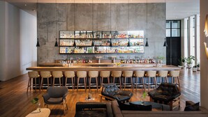 Uma área de bar moderna com balcão alto, diversas opções de assento incluindo banquetas e cadeiras estofadas, e variedade de garrafas e copos nas prateleiras.