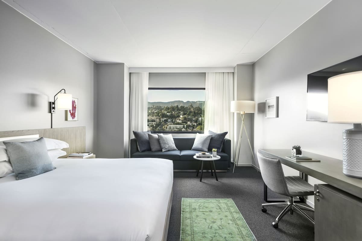 A hotel room with a bed, a sofa, a desk, and a chair. There is a window with a view of a cityscape.