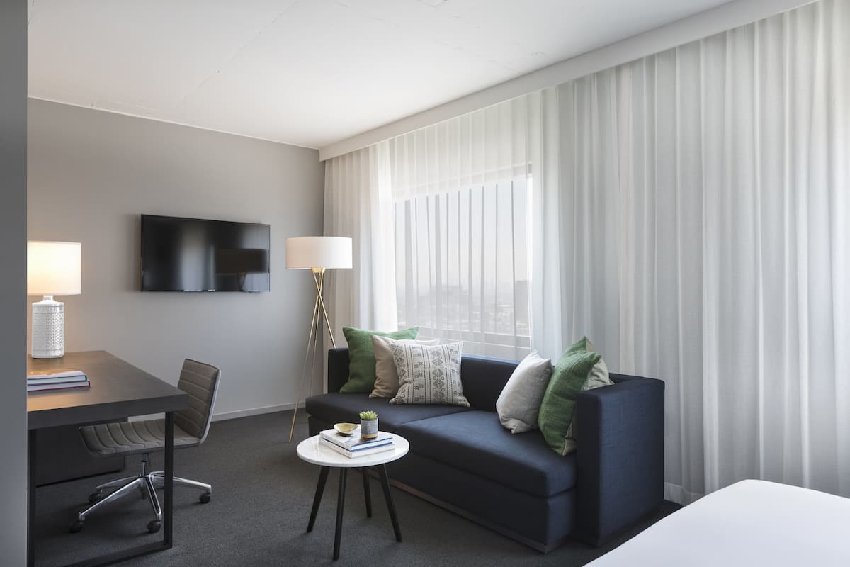 A modern hotel room with a dark blue sofa, a small round table with a lamp, a desk with a chair, and a flat-screen TV mounted on the wall.