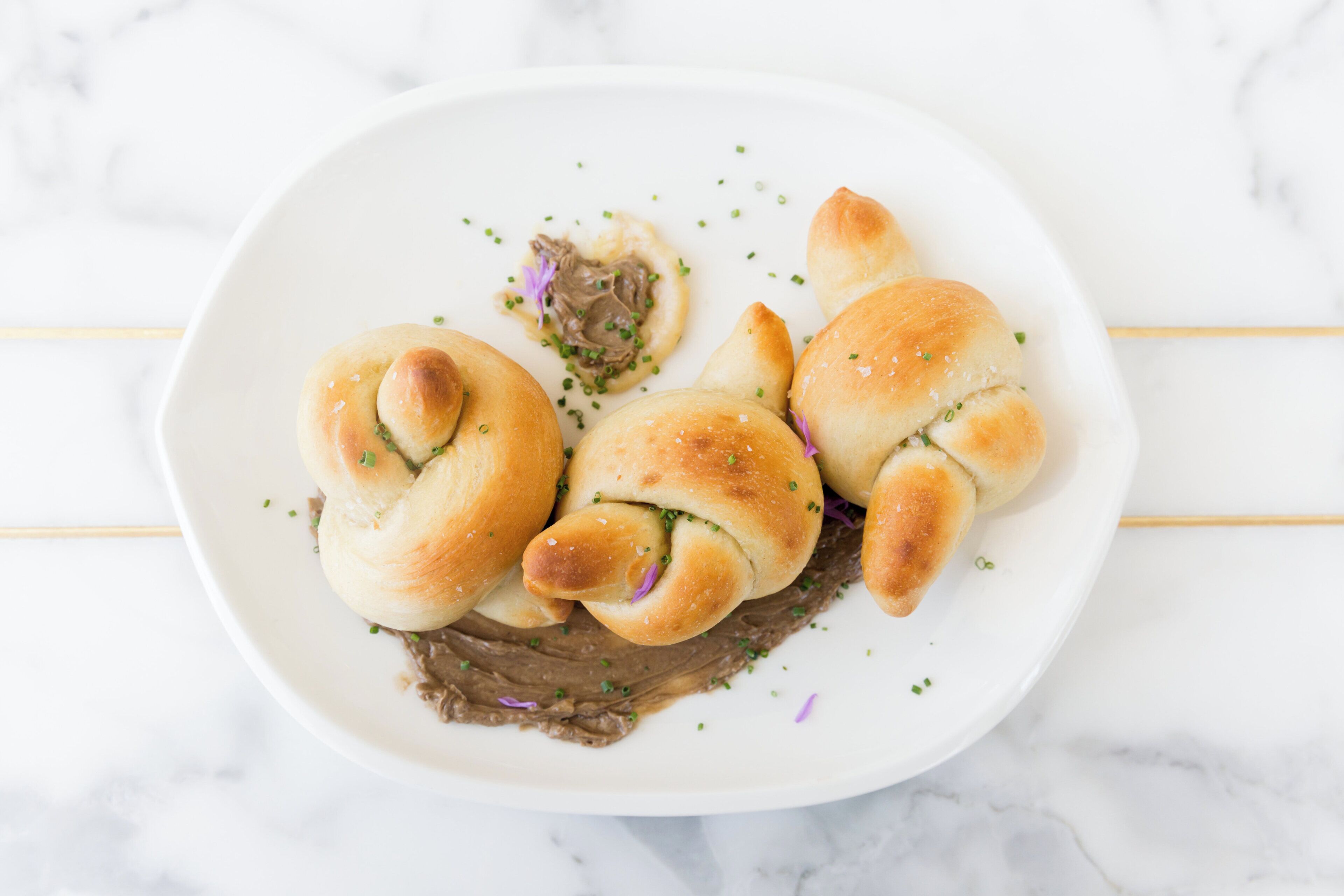 A white plate with three twisted rolls of bread, garnished with green herbs, on a marble surface.
