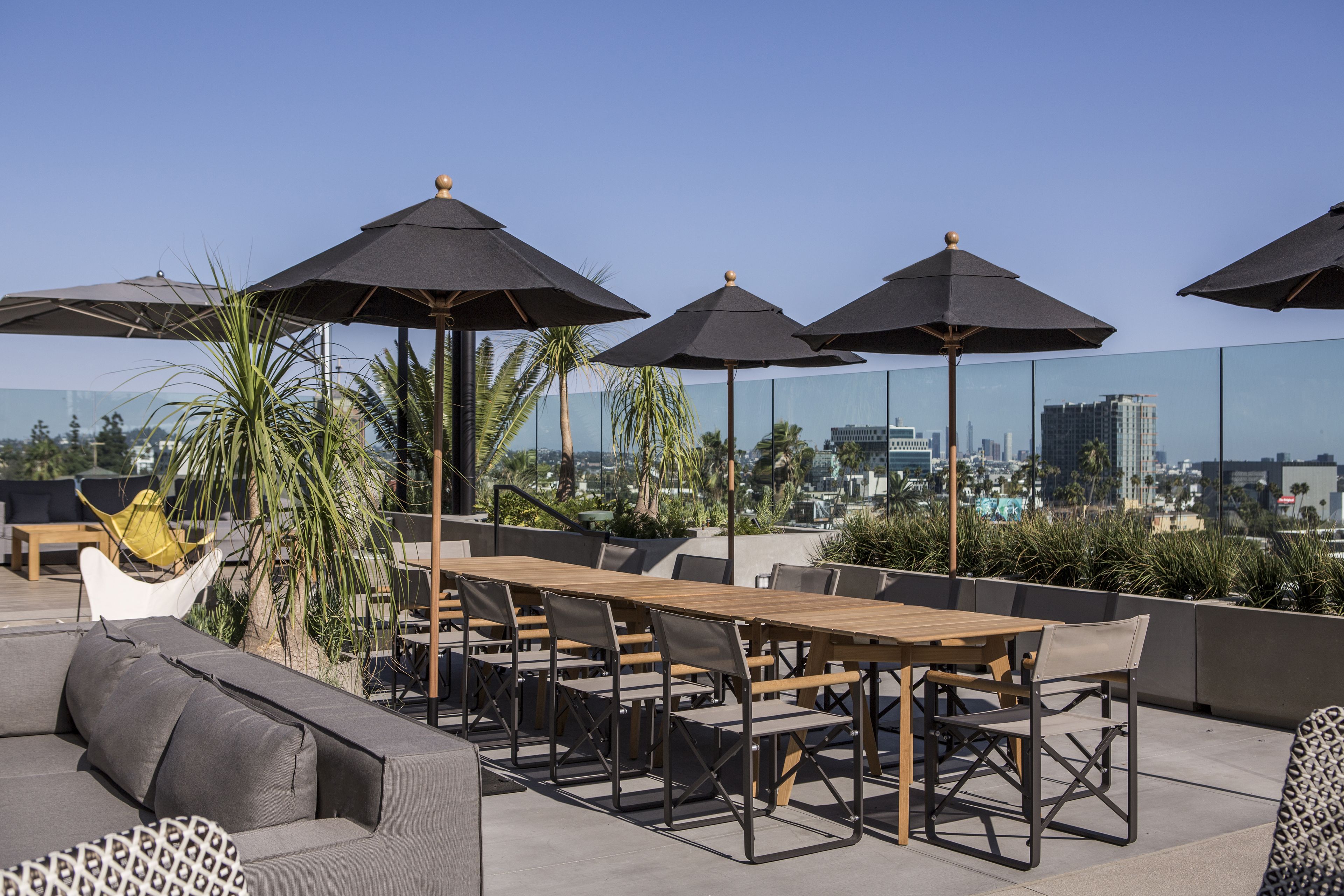 Eine Dachterrasse mit Holztisch und -stühlen, umgeben von schwarzen Sonnenschirmen, mit Blick auf die Stadt im Hintergrund.