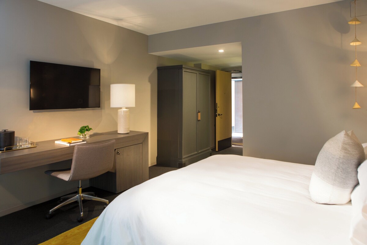Habitación de hotel con cama, escritorio, silla, televisión, armario y lámpara.
