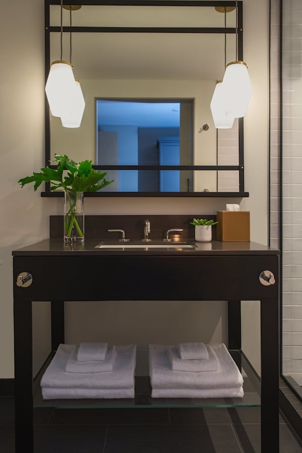 Un baño moderno con un tocador negro, un estante de vidrio con toallas dobladas, una planta en maceta y un espejo con luces colgantes.