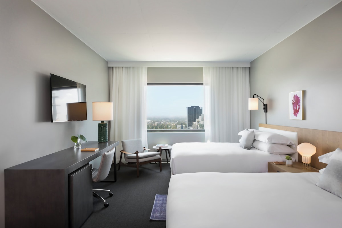 A hotel room with two beds, a desk, a chair, a small table, and a window with a city view.