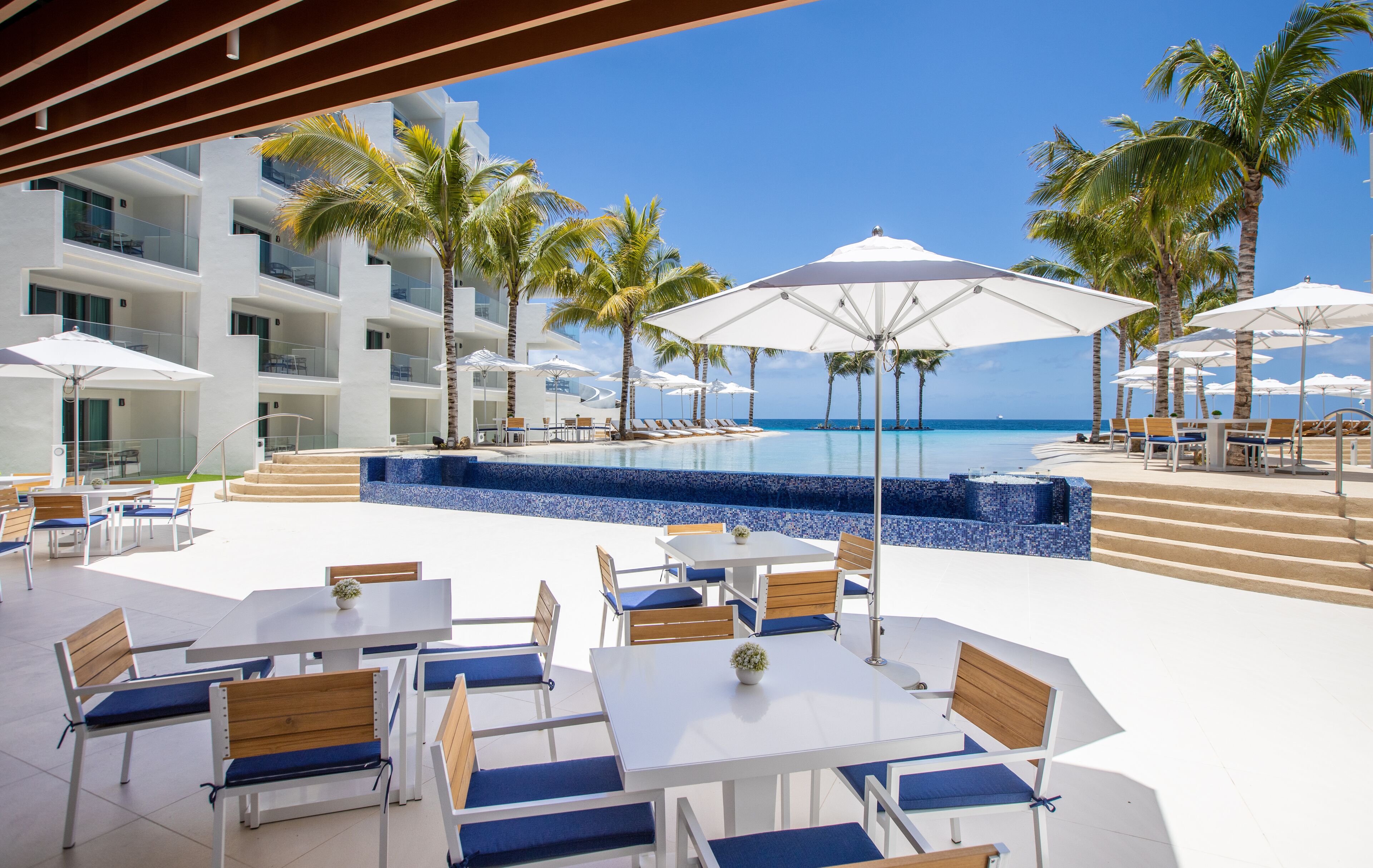 A resort pool area with multiple lounge chairs and tables, surrounded by palm trees and a clear view of the ocean.