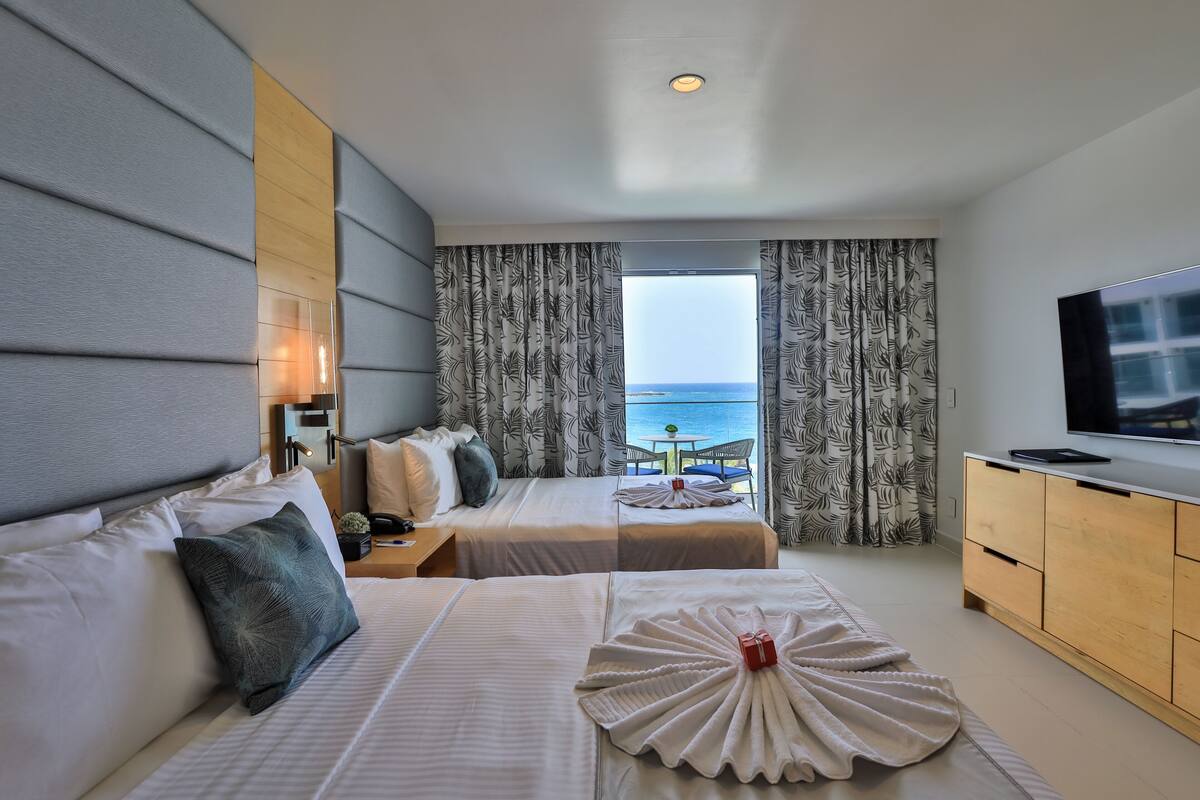 A hotel room with two beds, a TV, and a view of the sea.
