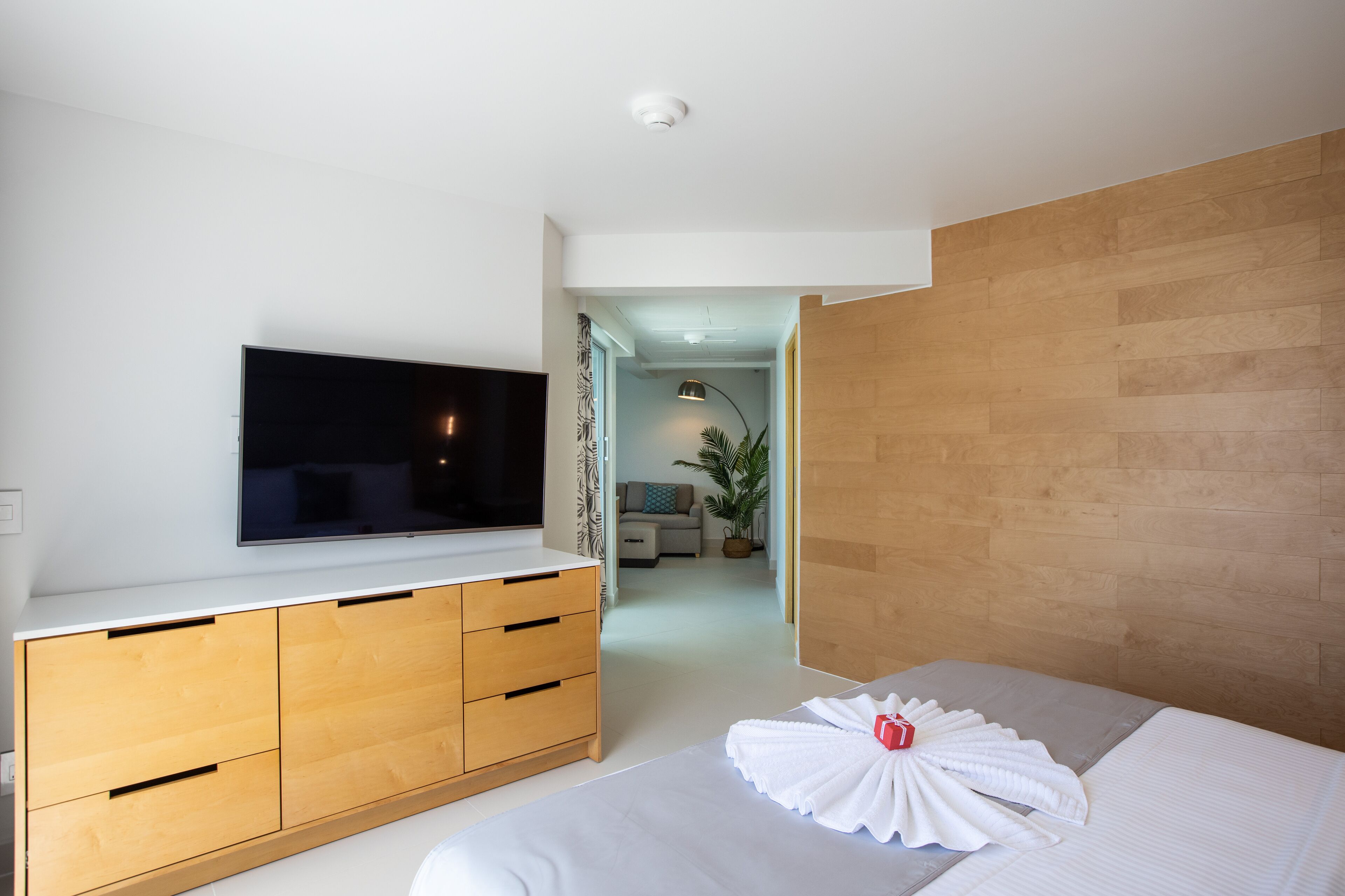 Une chambre d’hôtel moderne avec un téléviseur à écran plat fixé sur un mur blanc, une commode en bois avec des tiroirs, un lit soigneusement fait avec une serviette, et une vue sur le salon adjacent.
