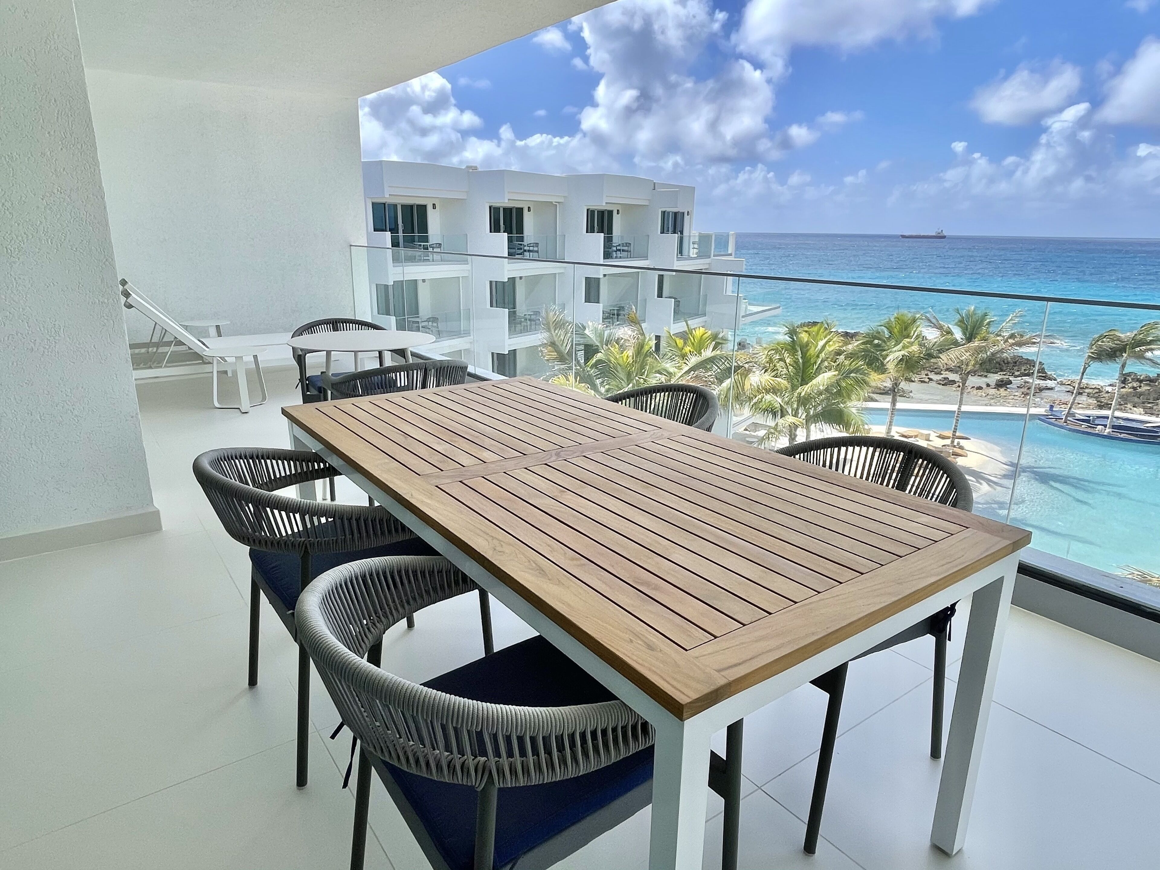 A balcony with a wooden table and chairs, overlooking a beach and ocean.