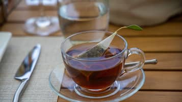 A clear glass cup of tea with a tea bag on a saucer, placed on a wooden table with a napkin and a silver spoon beside it.