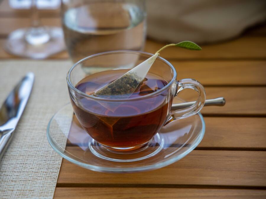 A clear glass cup of tea with a tea bag on a saucer, placed on a wooden table with a napkin and a silver spoon beside it.