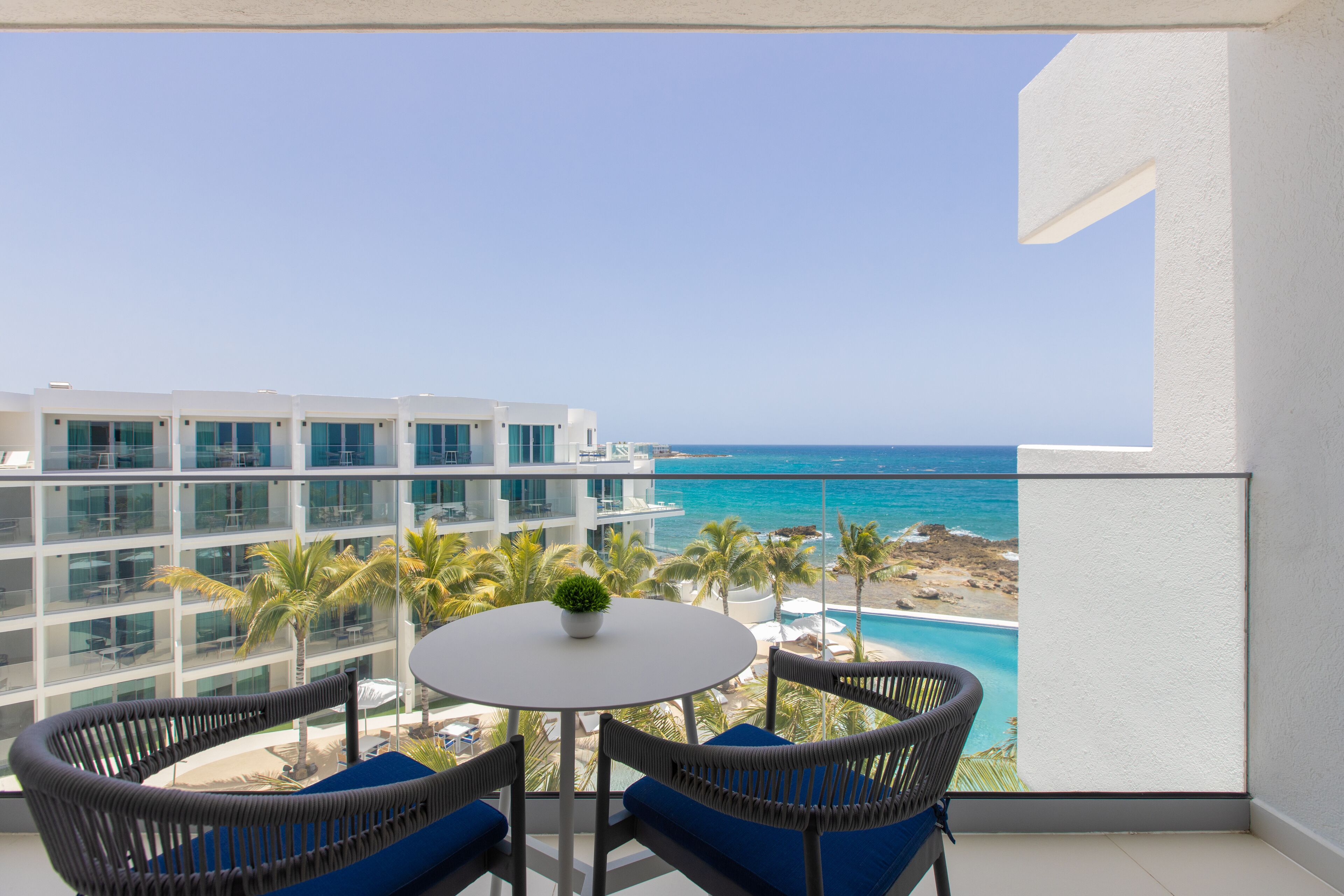A balcony with a round table and wicker chairs, overlooking a beach and ocean.