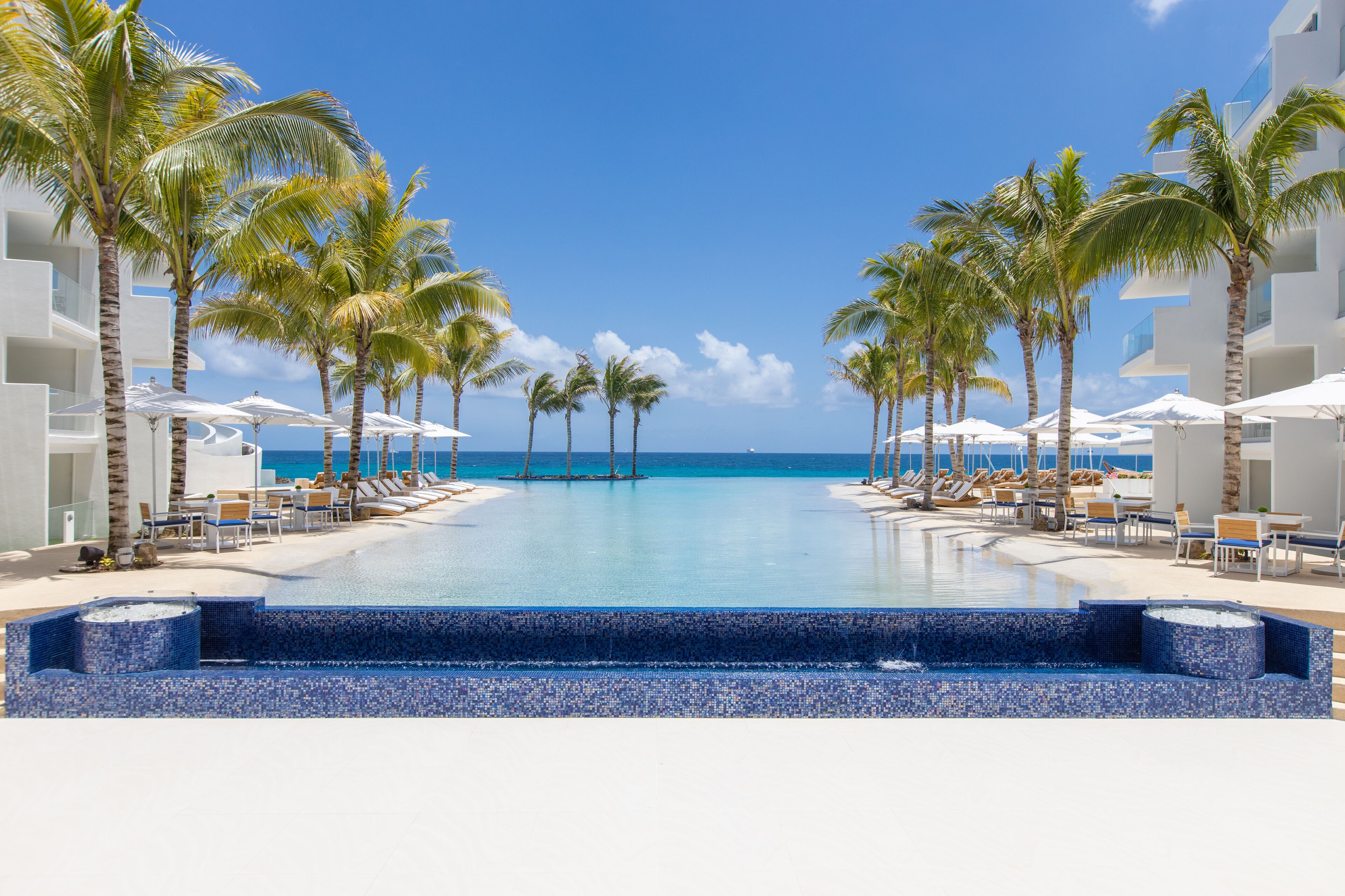 A poolside area with lounge chairs and umbrellas, palm trees, and a clear view of the ocean.