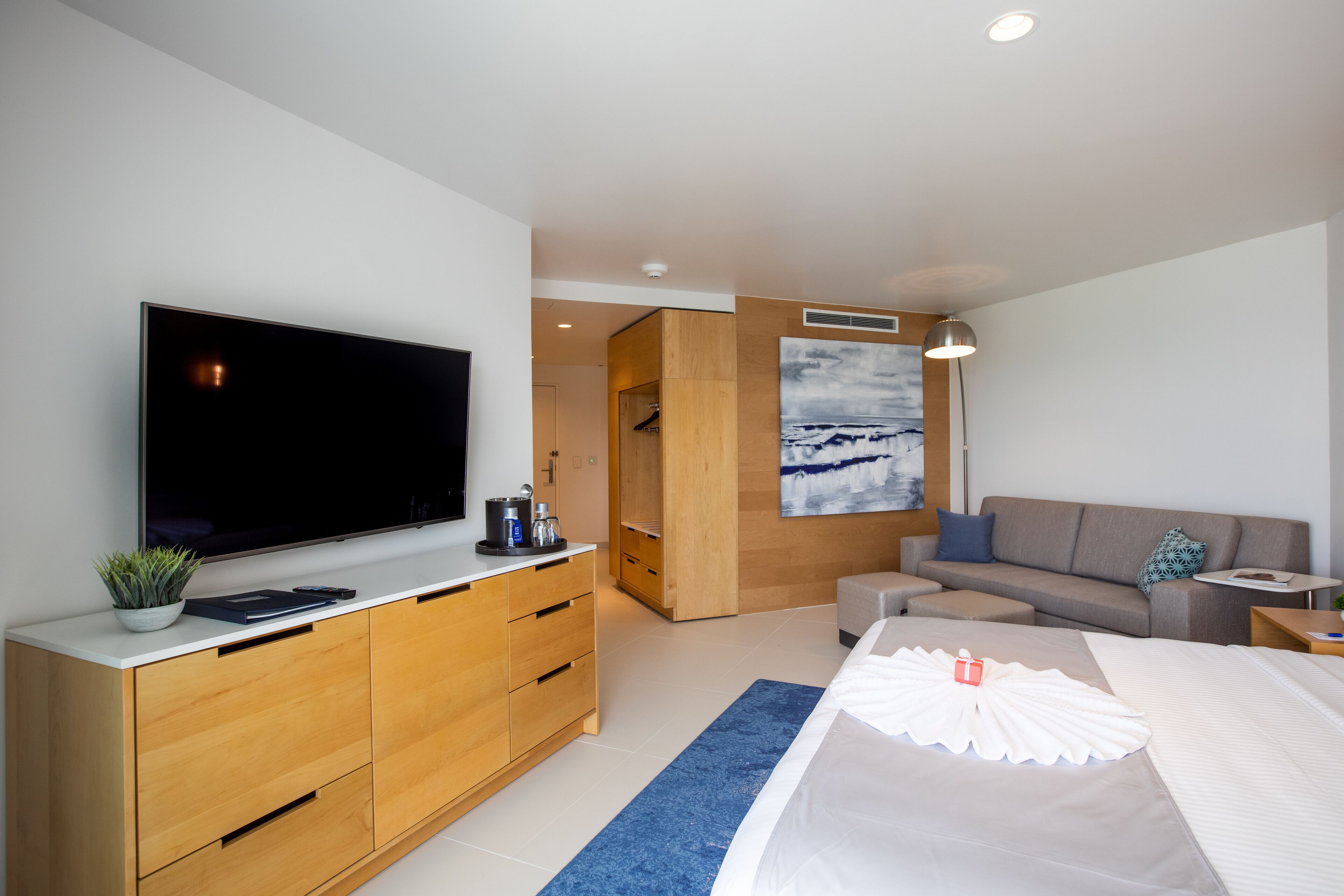 A modern hotel room with a large flat-screen TV, wooden furniture, a sofa, a bed with a white comforter, and a painting on the wall.