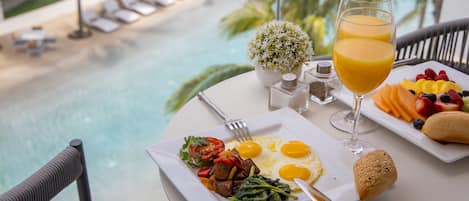 A breakfast table with plates of eggs, toast, and fruit, a glass of orange juice, and a view of a pool and lounge area.