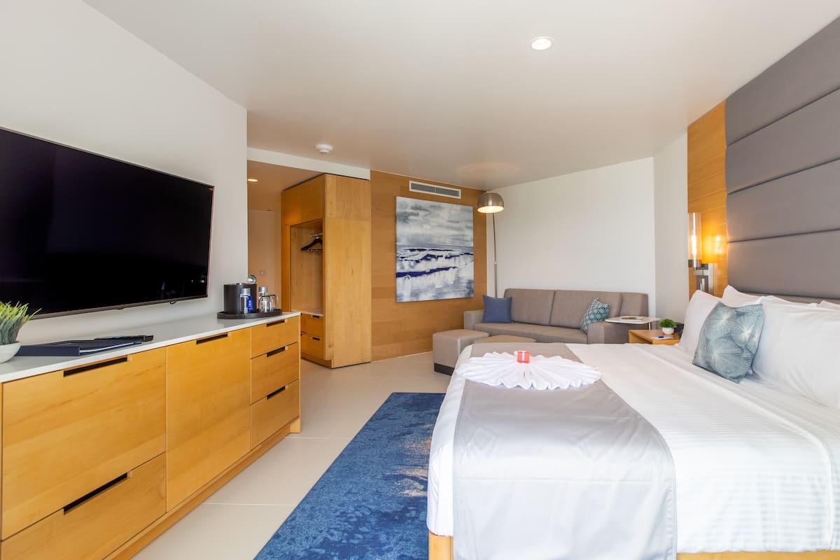 A modern hotel room with a large flat-screen TV, wooden furniture, a sofa, a bed with white bedding, and a blue rug.