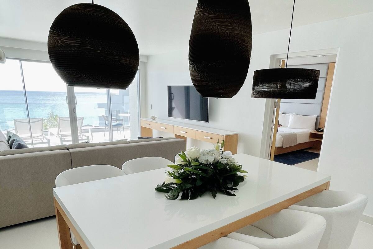 A modern dining area with a white table, four white chairs, and a floral centerpiece. A sofa and a TV are visible in the background.