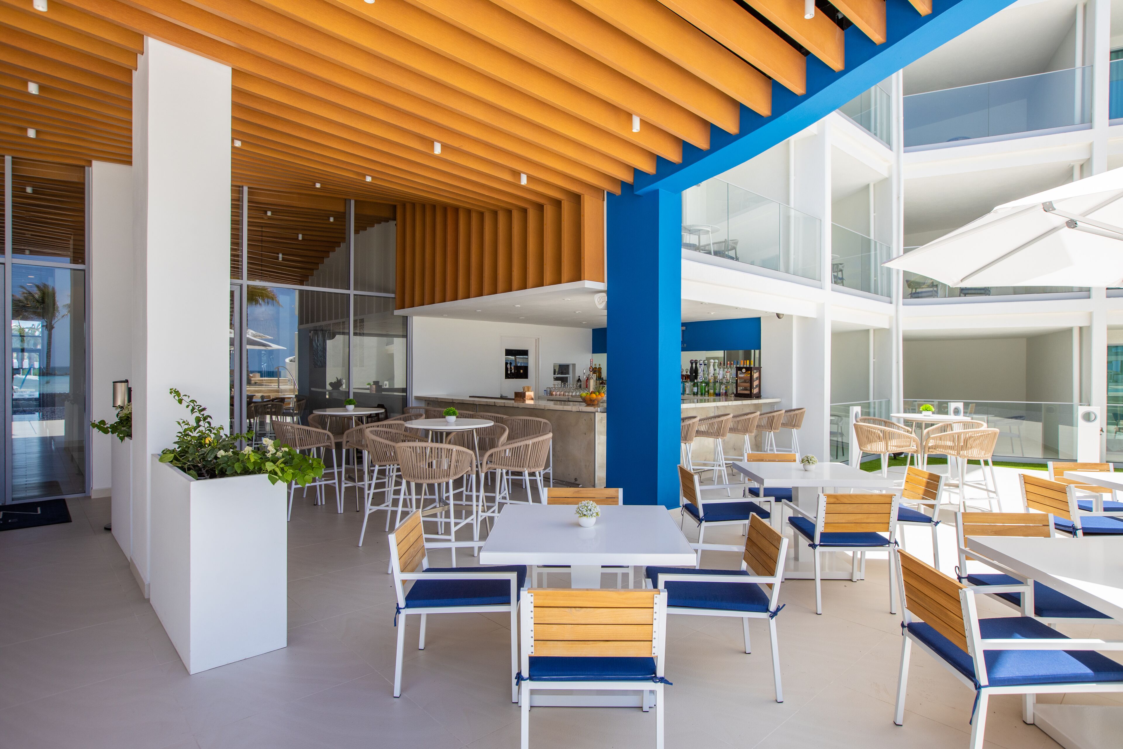 A modern indoor dining area with white tables and chairs, a wooden ceiling, and a glass partition separating the dining area from a kitchen.