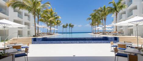 Zona de piscina en un resort con vista al océano, rodeada de palmeras y sillones de descanso.