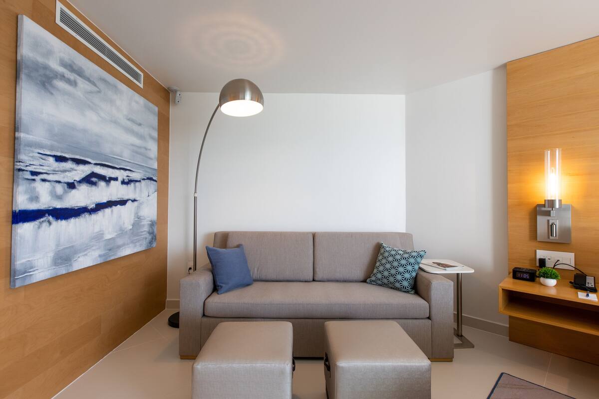 A modern living room with a grey sofa, a standing lamp, a coffee table, and a wall-mounted shelf with a plant and a clock.