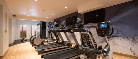Un gimnasio con varias cintas de correr y bicicletas estáticas, piso de madera y un gran mural en la pared.