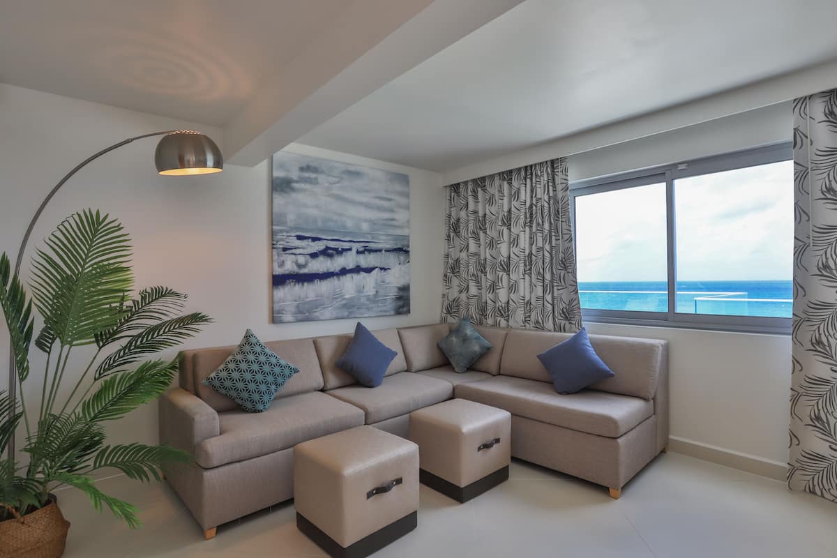 A modern living room with a sectional sofa, ottomans, a floor lamp, a large painting, and a view of the ocean through a window.