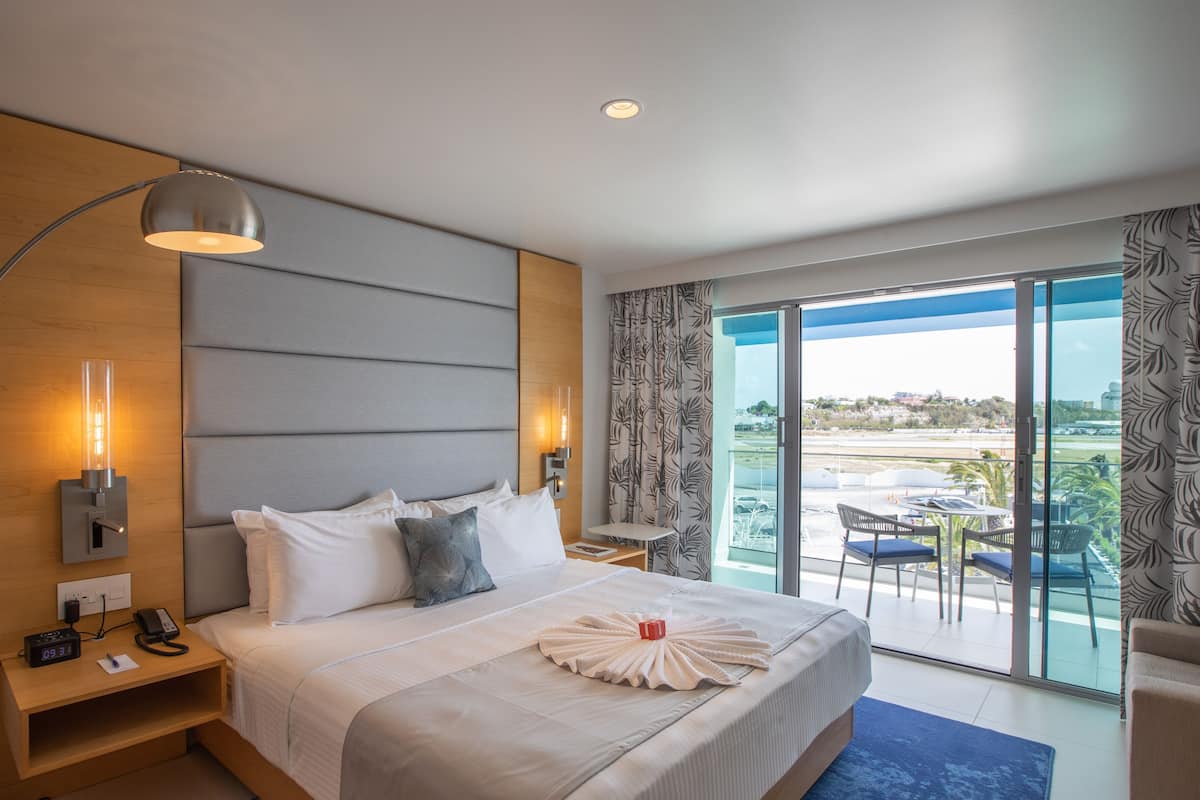 A hotel room with a large bed, a bedside table with a lamp, a telephone, and a clock. There is a view of a balcony with chairs and a table.