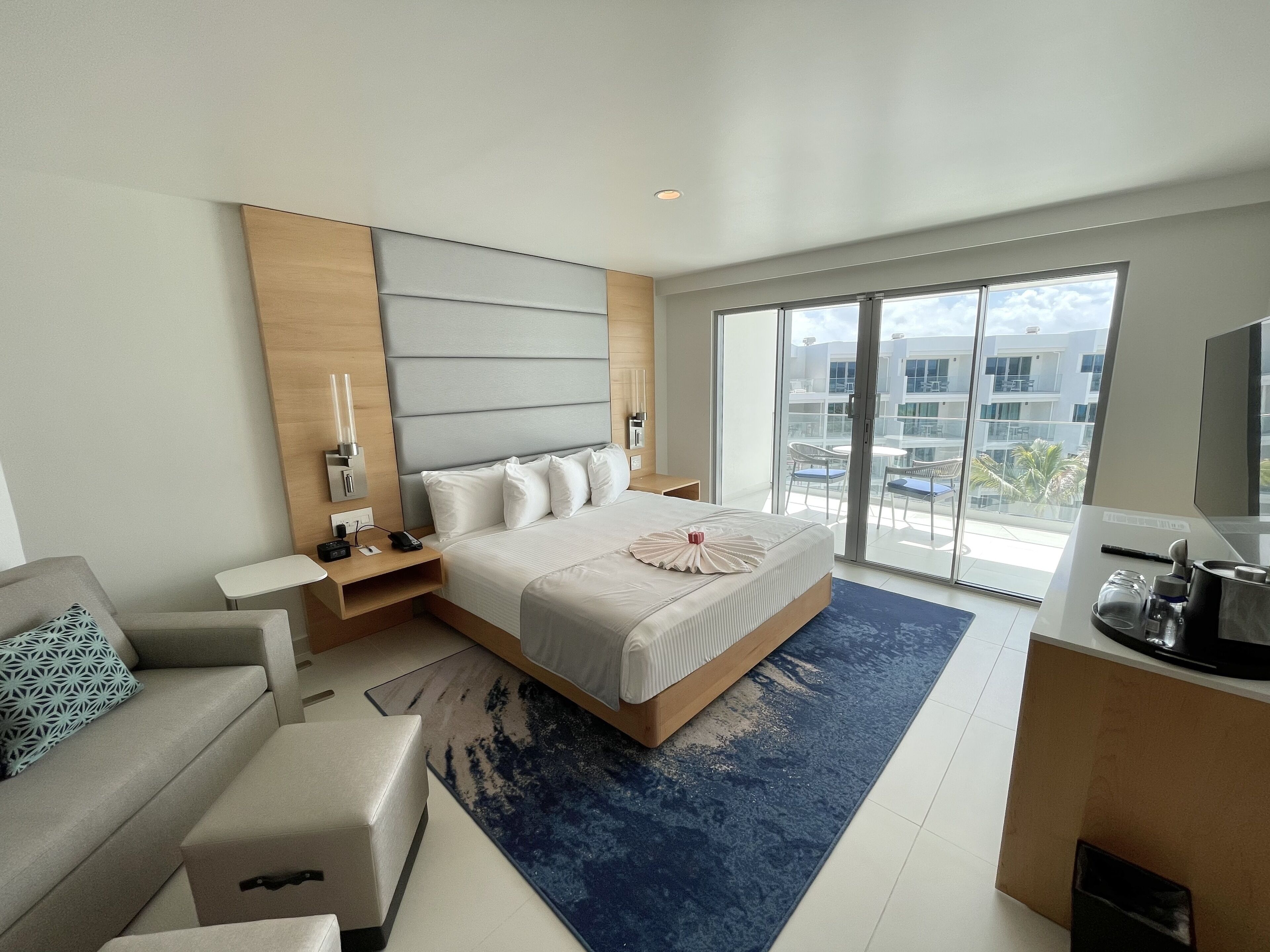 A modern hotel room with a large bed, a sofa, a desk, and a balcony with a city view.