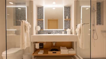Un baño moderno con ducha envuelta en vidrio, inodoro blanco, tocador con espejo y repisa con toallas.