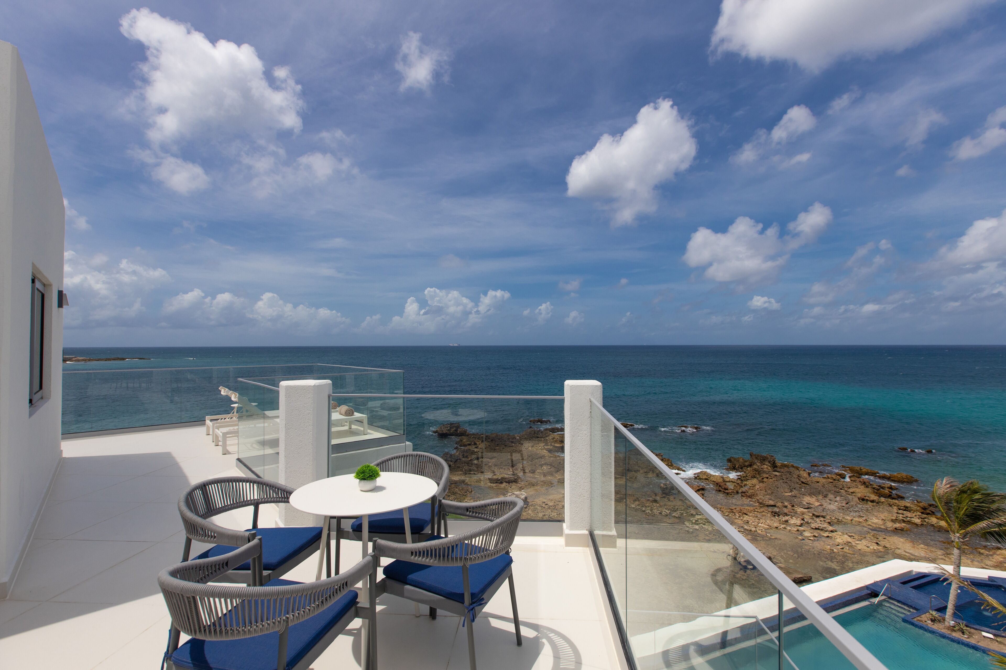 A balcony with a glass railing overlooking a beach and ocean, furnished with wicker chairs and a small table.