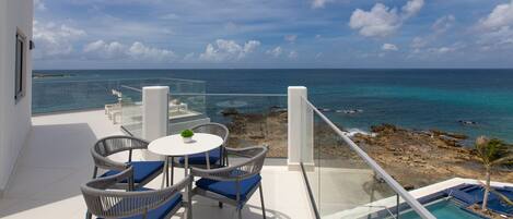 A balcony with a glass railing overlooking a beach and ocean, furnished with wicker chairs and a small table.