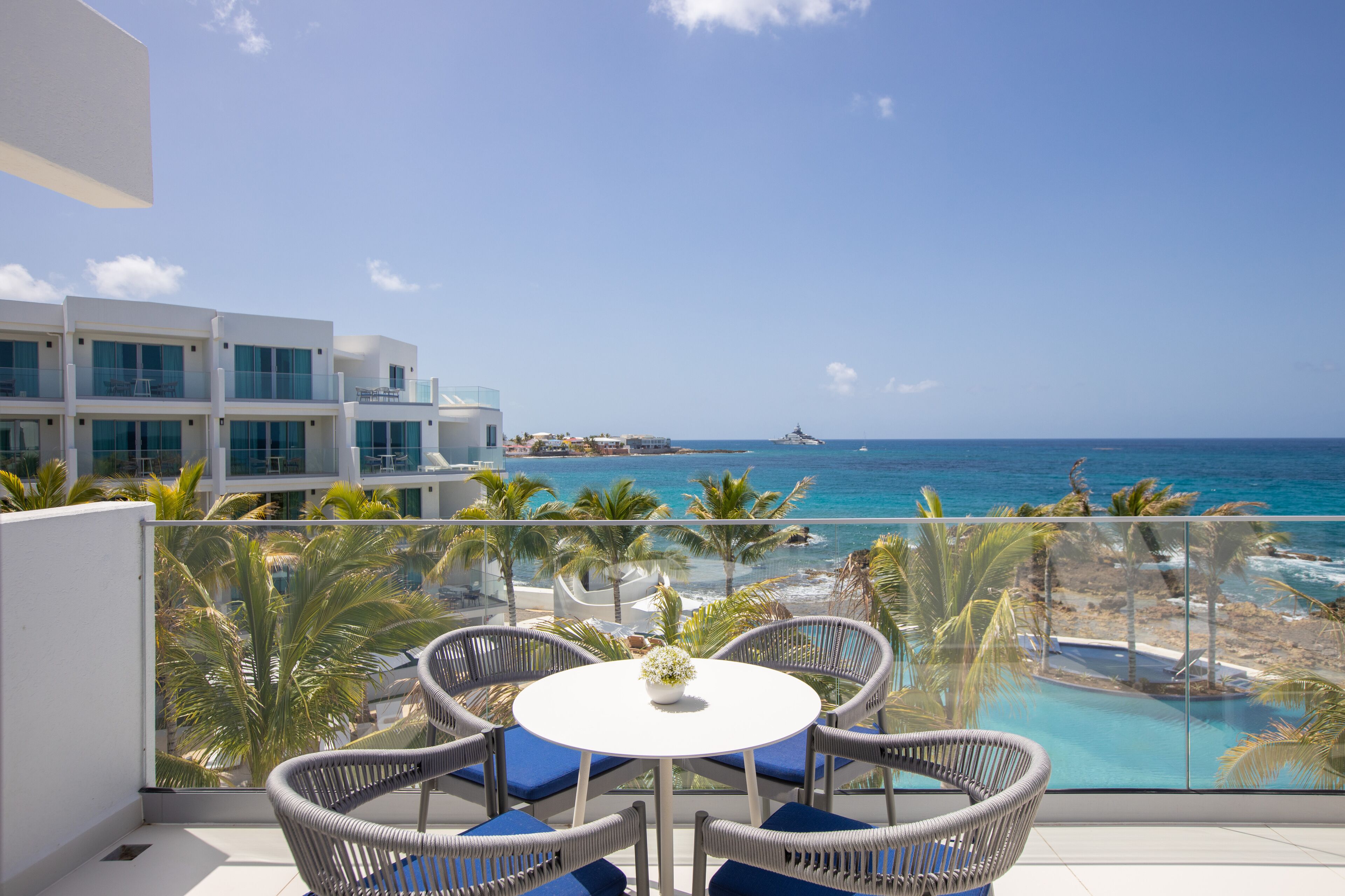 A balcony with wicker chairs and a table, overlooking a beach and ocean.