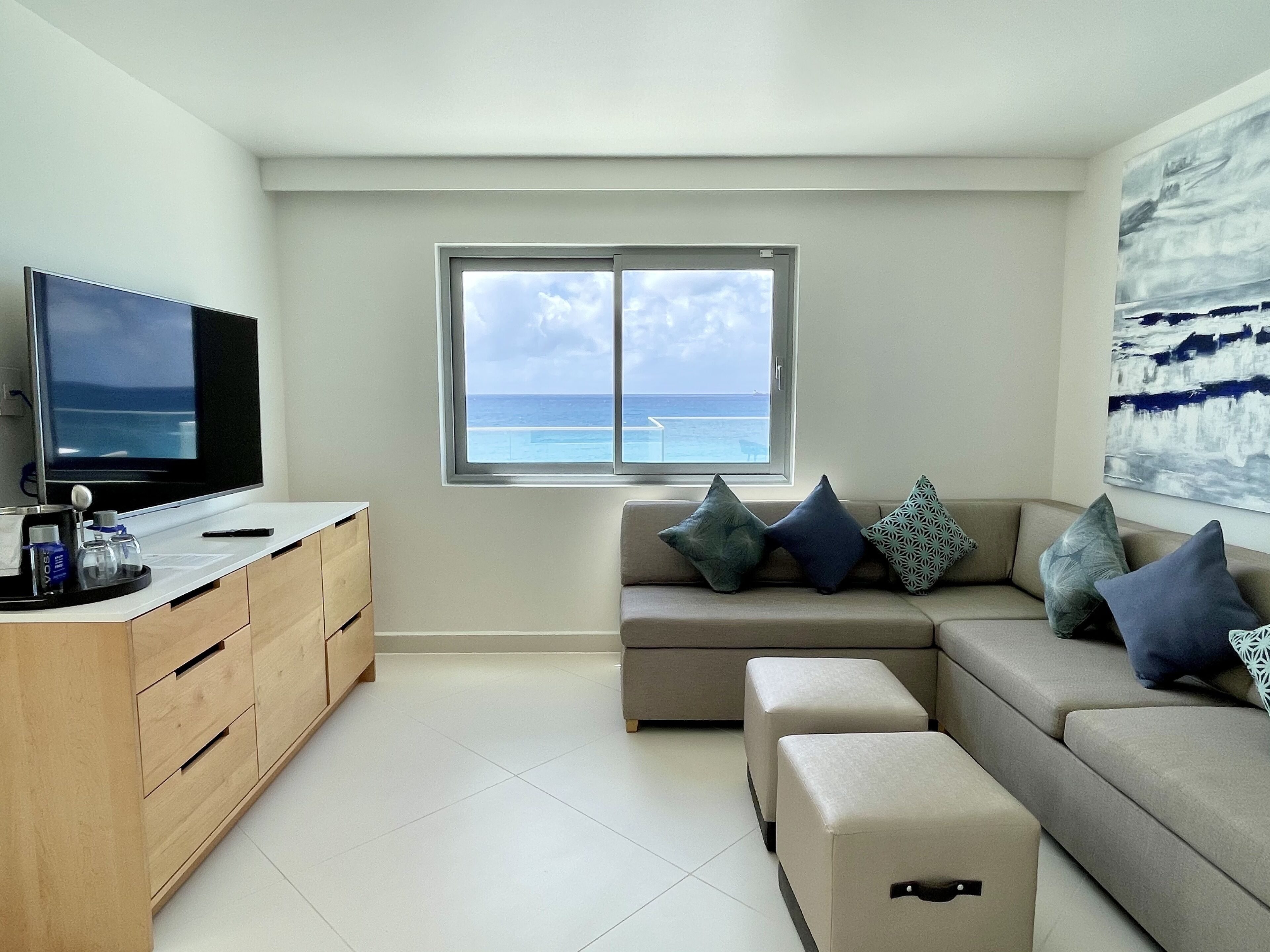 A modern living room with a large window offering a view of the ocean, a flat-screen TV, a wooden cabinet, and a beige sofa with patterned cushions.