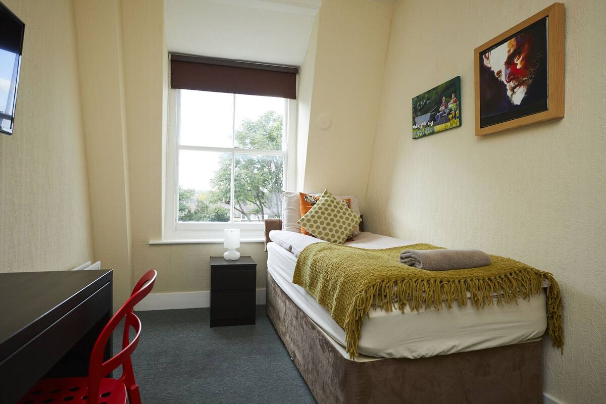 A bedroom with a bed, a desk, a chair, a window, and two paintings on the wall.