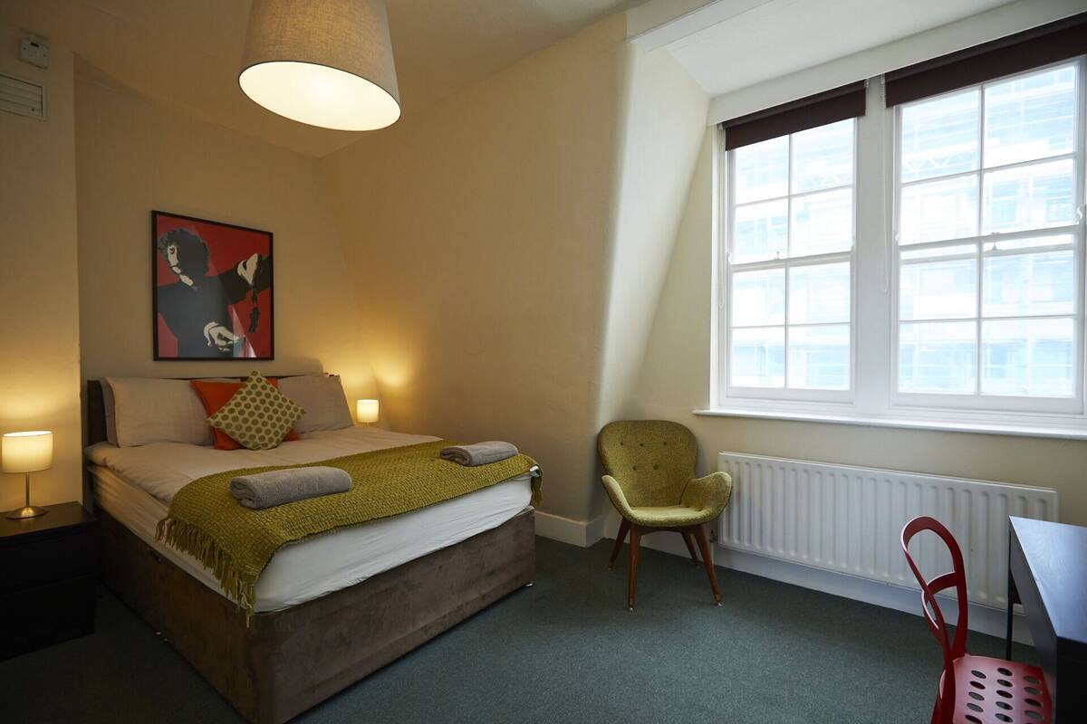 A hotel room with a bed, a chair, a table, a window, and a painting on the wall.